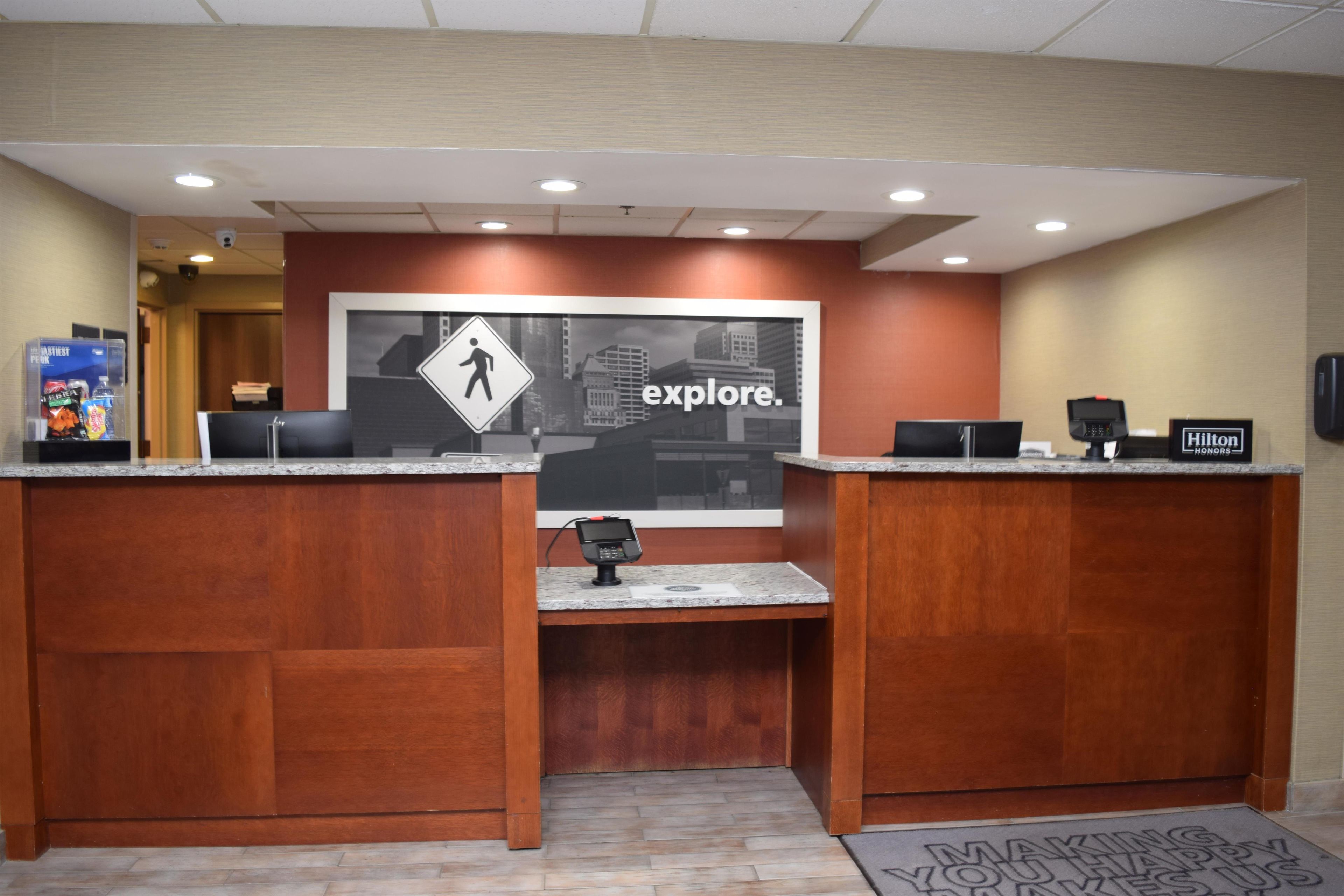 Un mostrador de recepción de hotel con un cartel grande que dice 'explore'. Hay dos mostradores de recepción, una máquina expendedora y un cartel de 'Hilton Honors'.