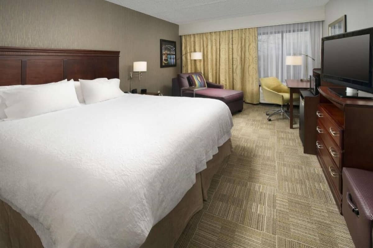 A hotel room with a large bed, a desk with a television, a chair, and a desk lamp.