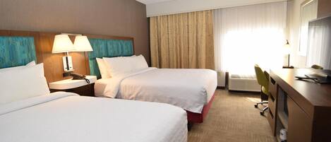 A hotel room with two beds, a desk, a chair, a lamp, and a window with curtains.