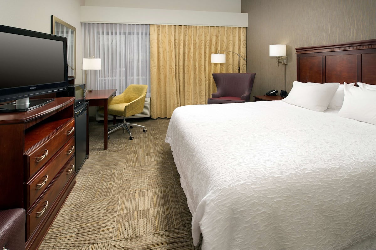 A hotel room with a large bed, a television, a desk with a chair, and a lamp.