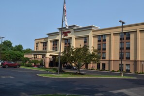 Un hôtel Hampton Inn avec un mât de drapeau, un parking et un arbre devant l’entrée.