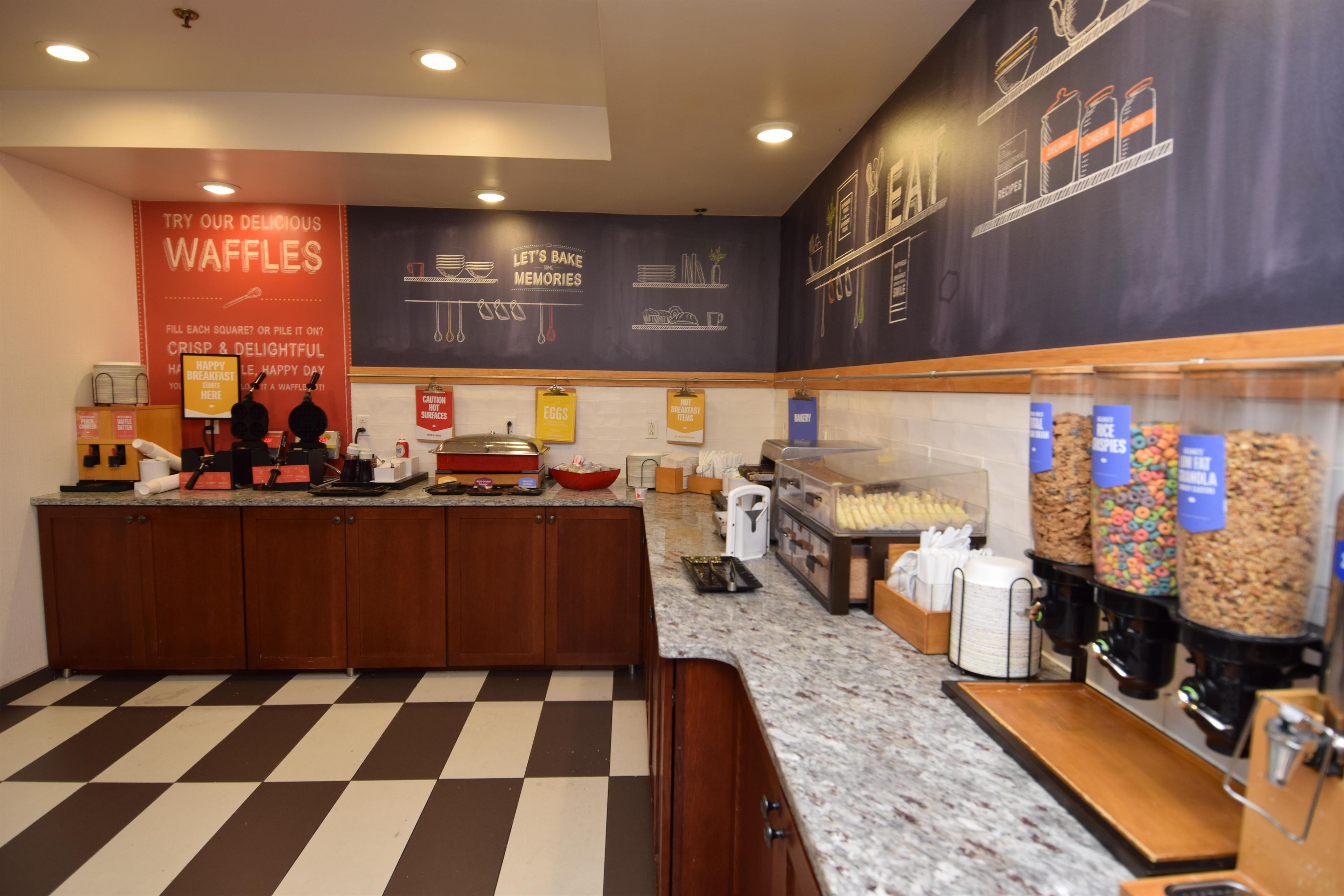A breakfast area with waffle-making equipment, a coffee station, and a wall with chalkboard-style menu writing.