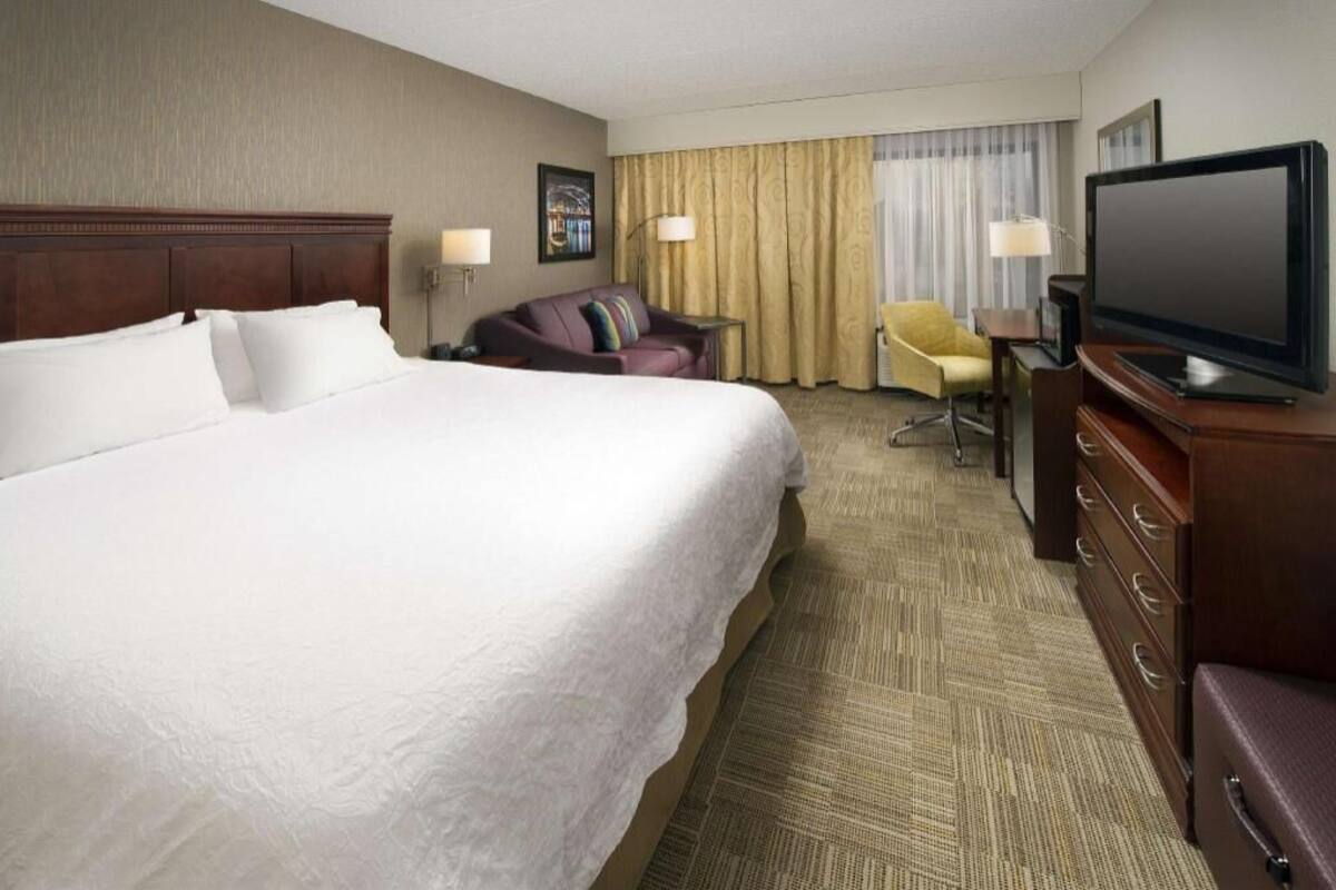 A hotel room with a large bed, a television, a desk with a chair, and a window with curtains.