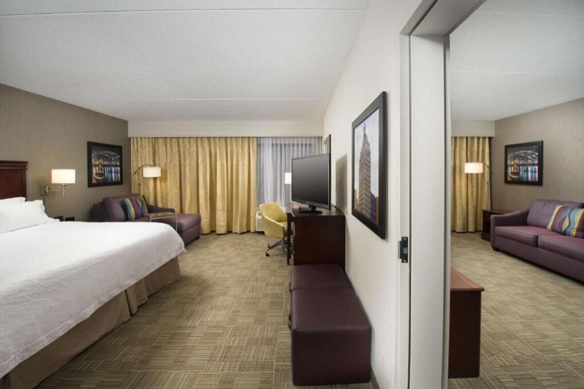A hotel room with a bed, a sofa, a desk with a chair, a television, and framed pictures on the walls.