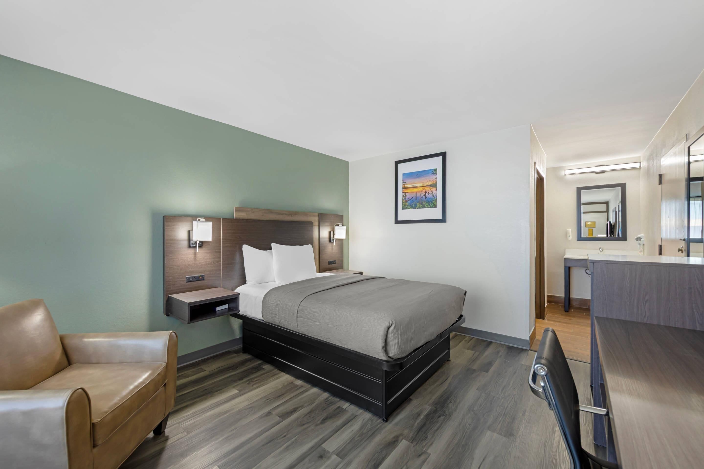 Une chambre d’hôtel avec un lit, une chaise, une table de chevet, une commode, un tableau et une salle de bain visible à travers une porte ouverte.
