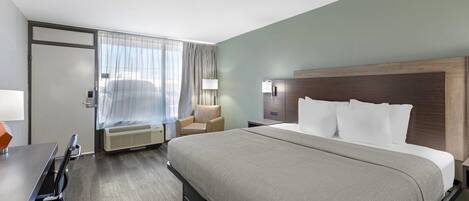 Una habitación de hotel con cama, escritorio, silla, lámpara y ventana con cortinas.