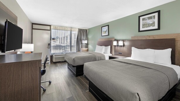 Ein Hotelzimmer mit zwei Betten, einem Schreibtisch, einem Fernseher und einem großen Fenster mit Vorhängen.