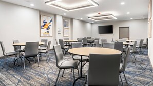 Une salle de conférence avec des tables et des chaises rondes, un téléviseur à écran plat fixé au mur et une moquette à motifs.