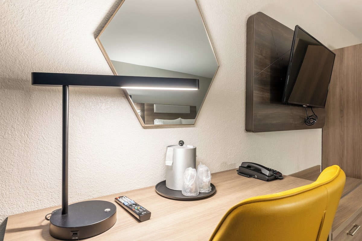 Habitación de hotel con escritorio, lámpara moderna, cafetera, teléfono y televisor montado en la pared.