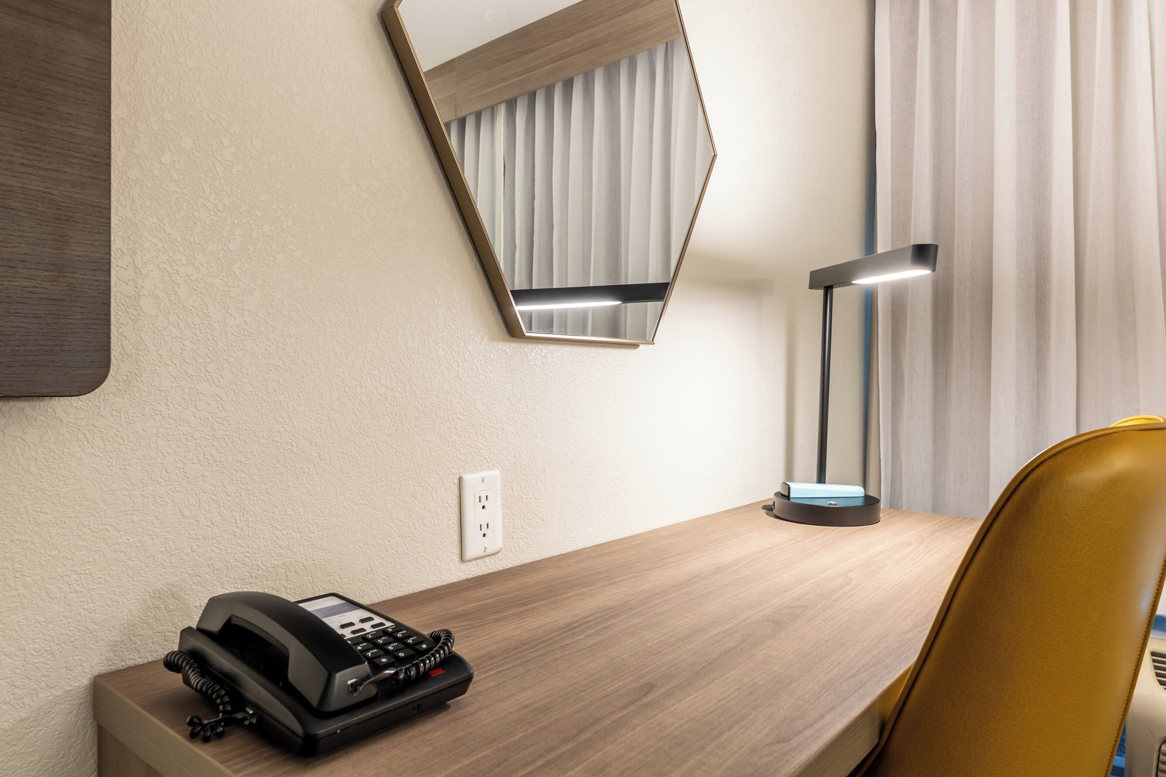 A hotel room with a wooden desk, a telephone, a mirror, a lamp, and a chair.