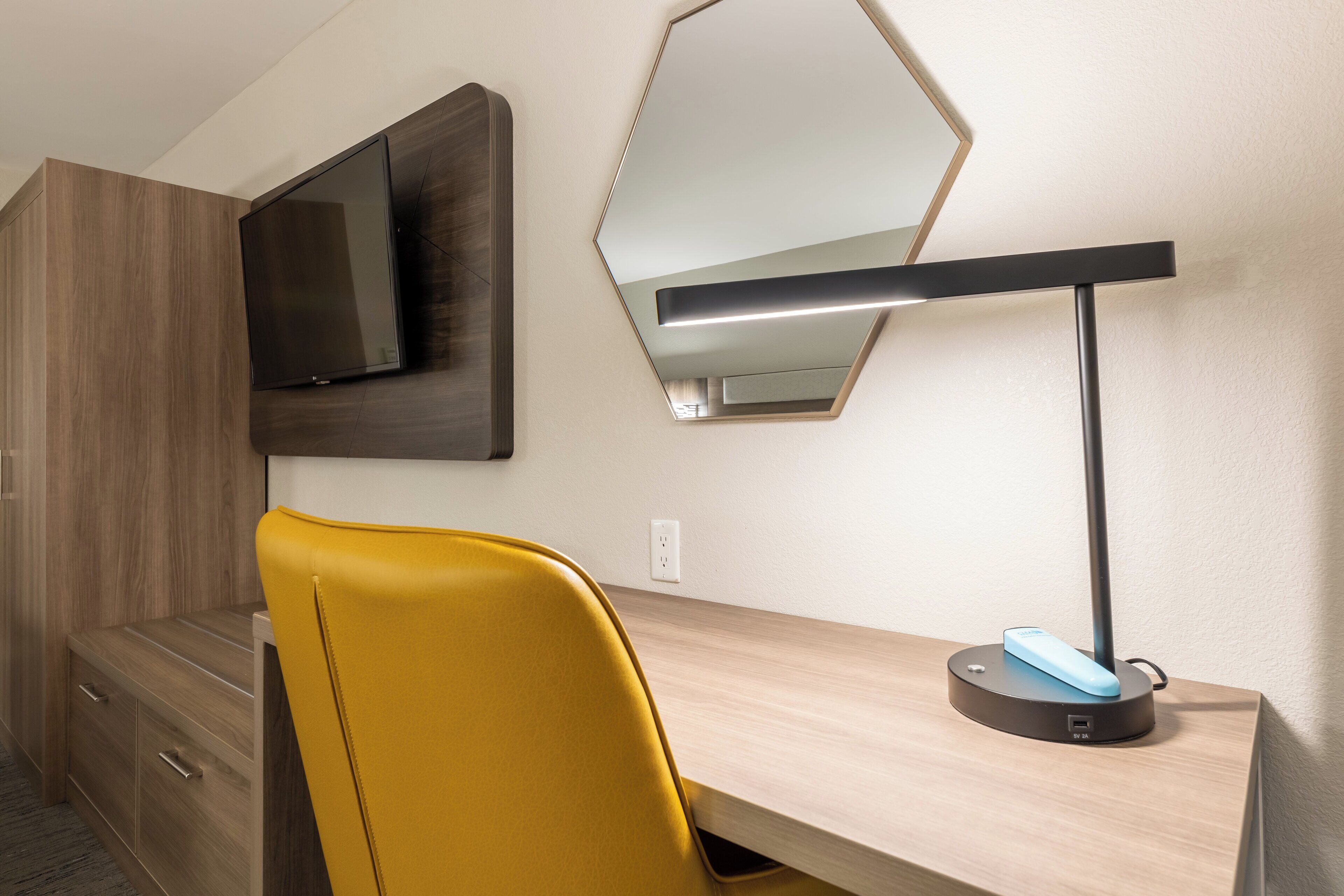 A modern hotel room with a wooden desk, a yellow chair, a flat-screen TV mounted on the wall, and a bedside table with a lamp.