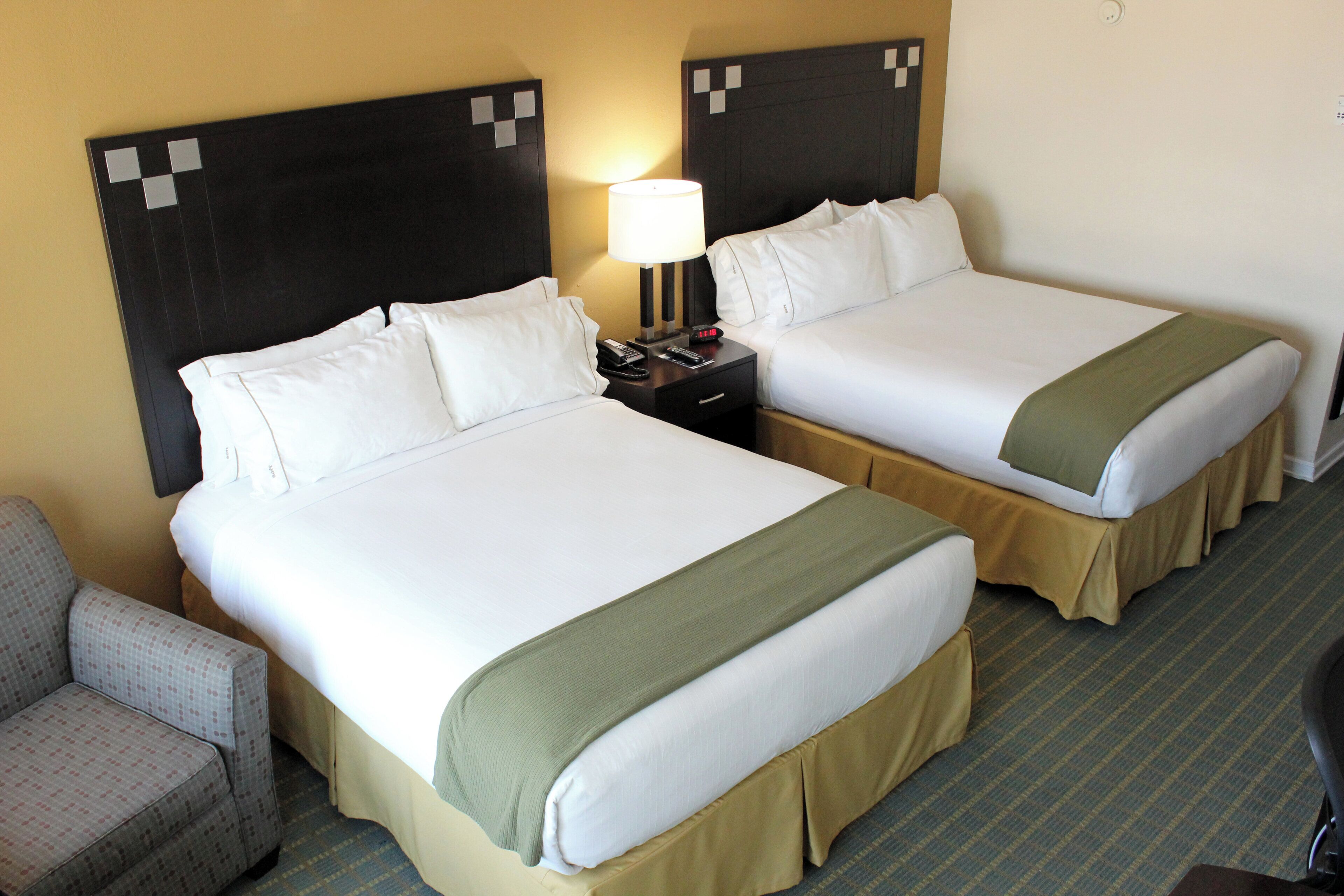 Habitación de hotel con dos camas, una mesita de noche con lámpara, una silla y piso alfombrado.