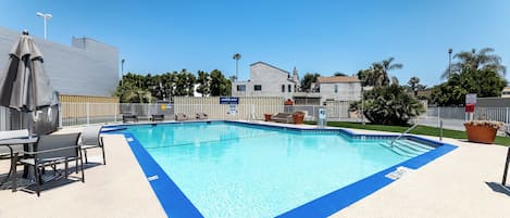 Una piscina rectangular con borde azul, rodeada por una terraza blanca. Se observan sillas de descanso y una mesa con sombrillas.
