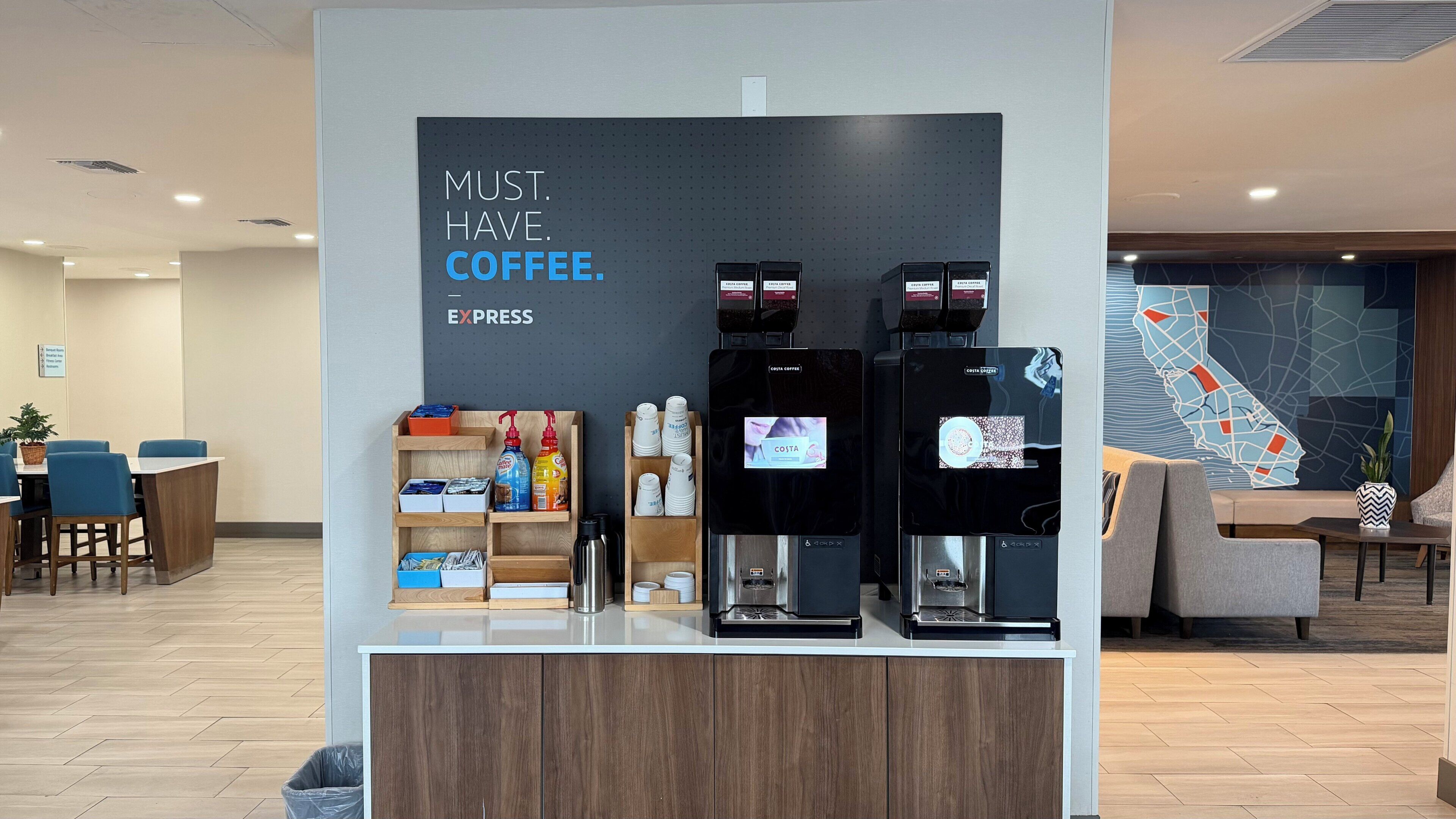 Una estación de Costa Coffee con dos máquinas de espresso, un dispensador de café y un panel informativo.