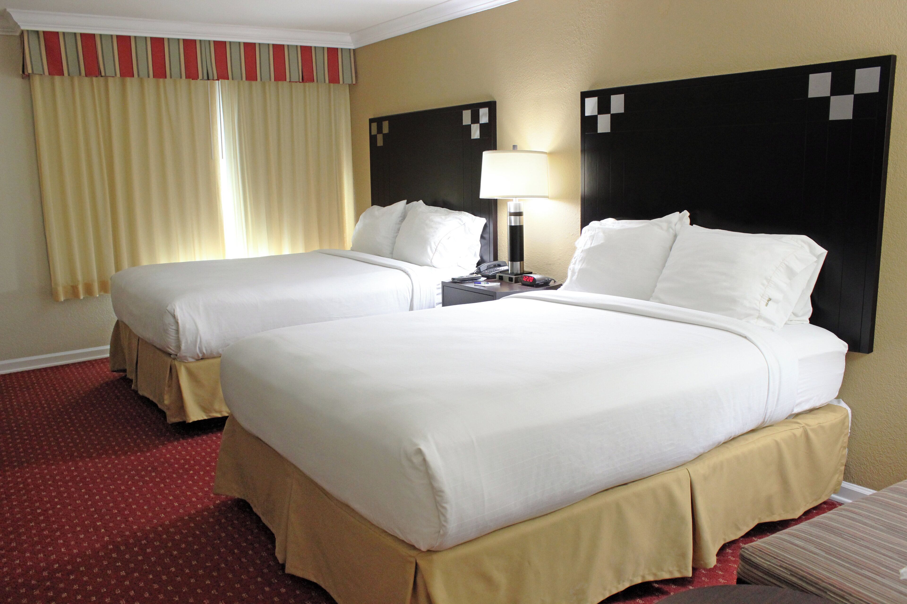 A hotel room with two beds, a nightstand, a lamp, and a window with curtains.