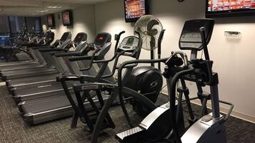 Un gimnasio con cintas de correr y máquinas elípticas, un ventilador y una pantalla digital que muestra listas de reproducción musical.