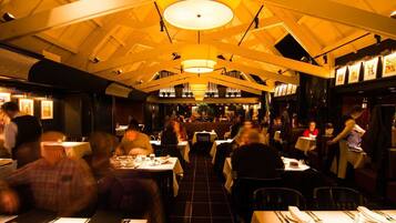 Restaurante com pé direito alto e iluminação acolhedora, com mesas e cadeiras dispostas para refeições.