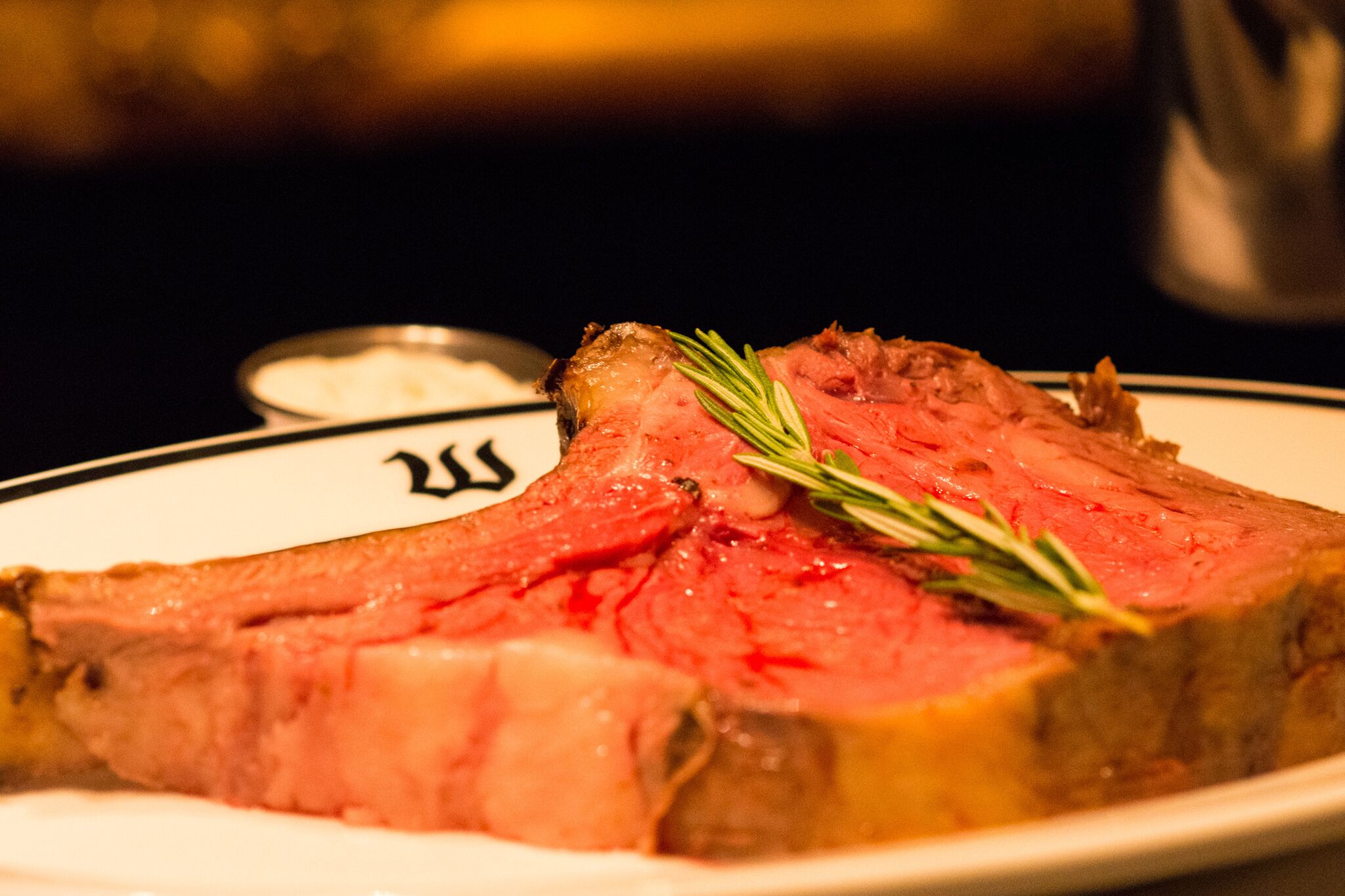A plate with a steak, a sprig of rosemary, and a small bowl of sauce.