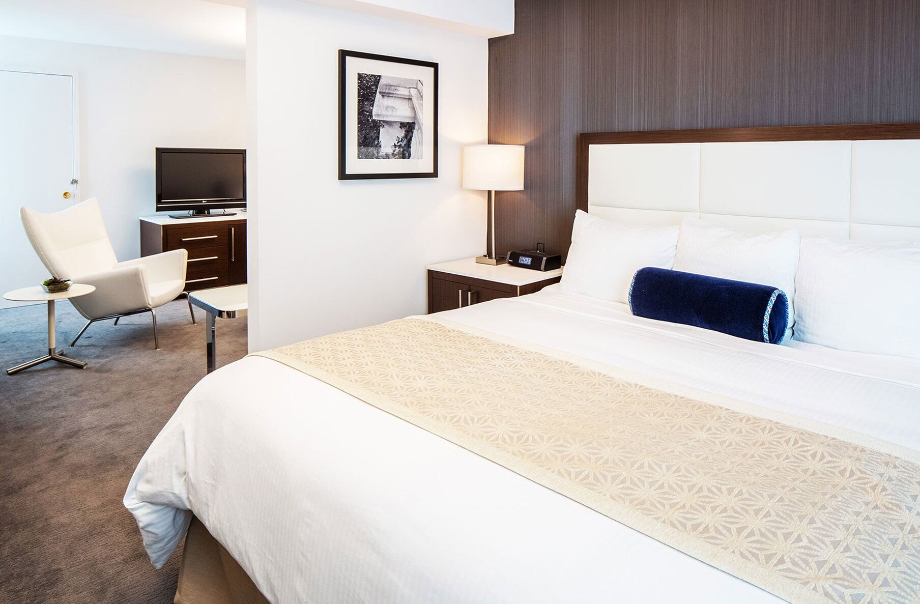 Ein Hotelzimmer mit einem großen Bett, einem Schreibtisch mit Computer, einem Sessel, einem Nachttisch mit Lampe und einem gerahmten Bild an der Wand.