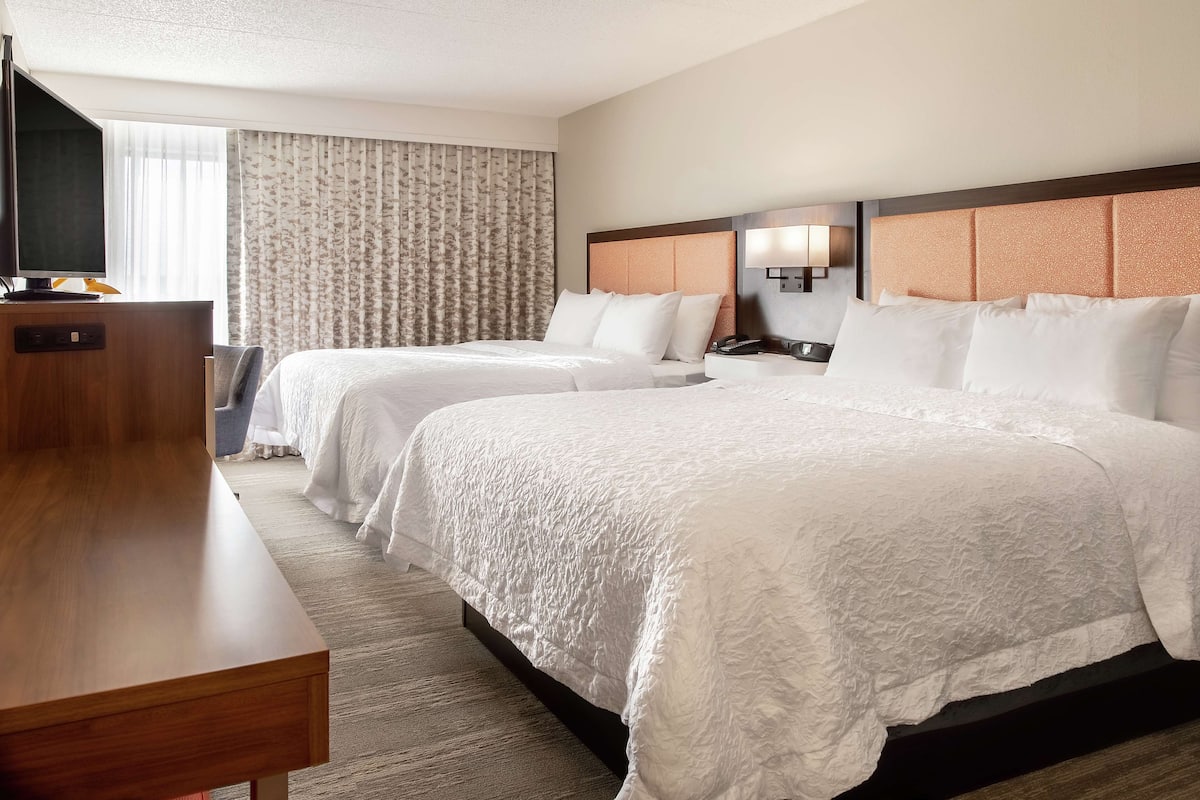 A hotel room with two beds, a wooden desk, a television, and a window with curtains.