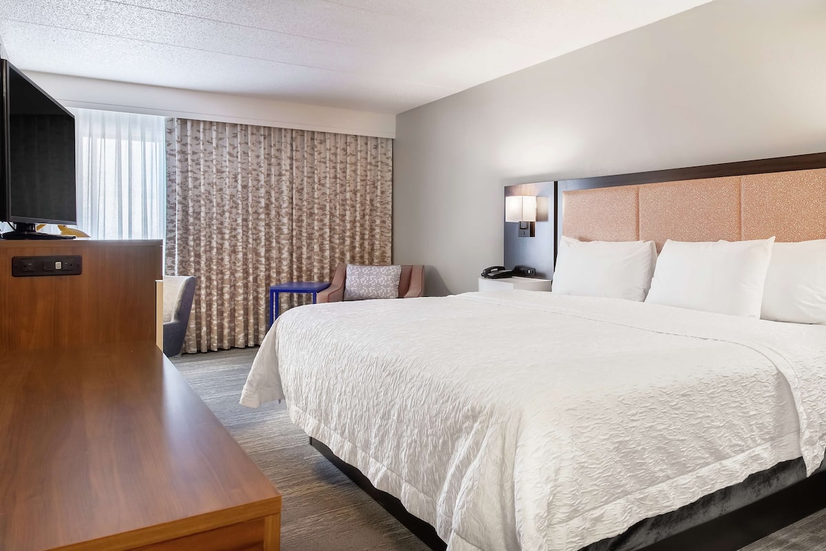 A hotel room with a large bed, a desk, a chair, a TV, and a window with curtains.