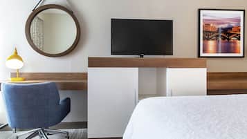 Habitación de hotel con cama, escritorio con televisor, silla, espejo redondo y un cuadro en la pared.
