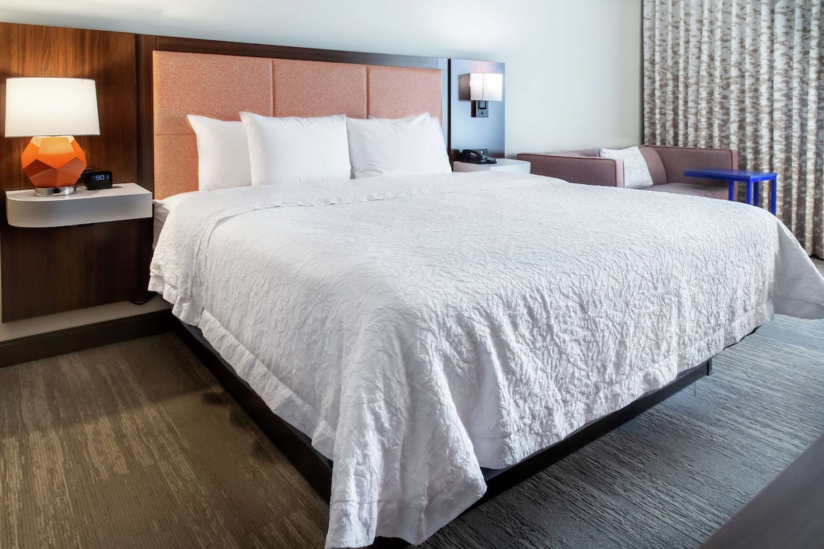A hotel room with a large bed, a nightstand with a lamp, a wall clock, a seating area with a table, and a curtain.