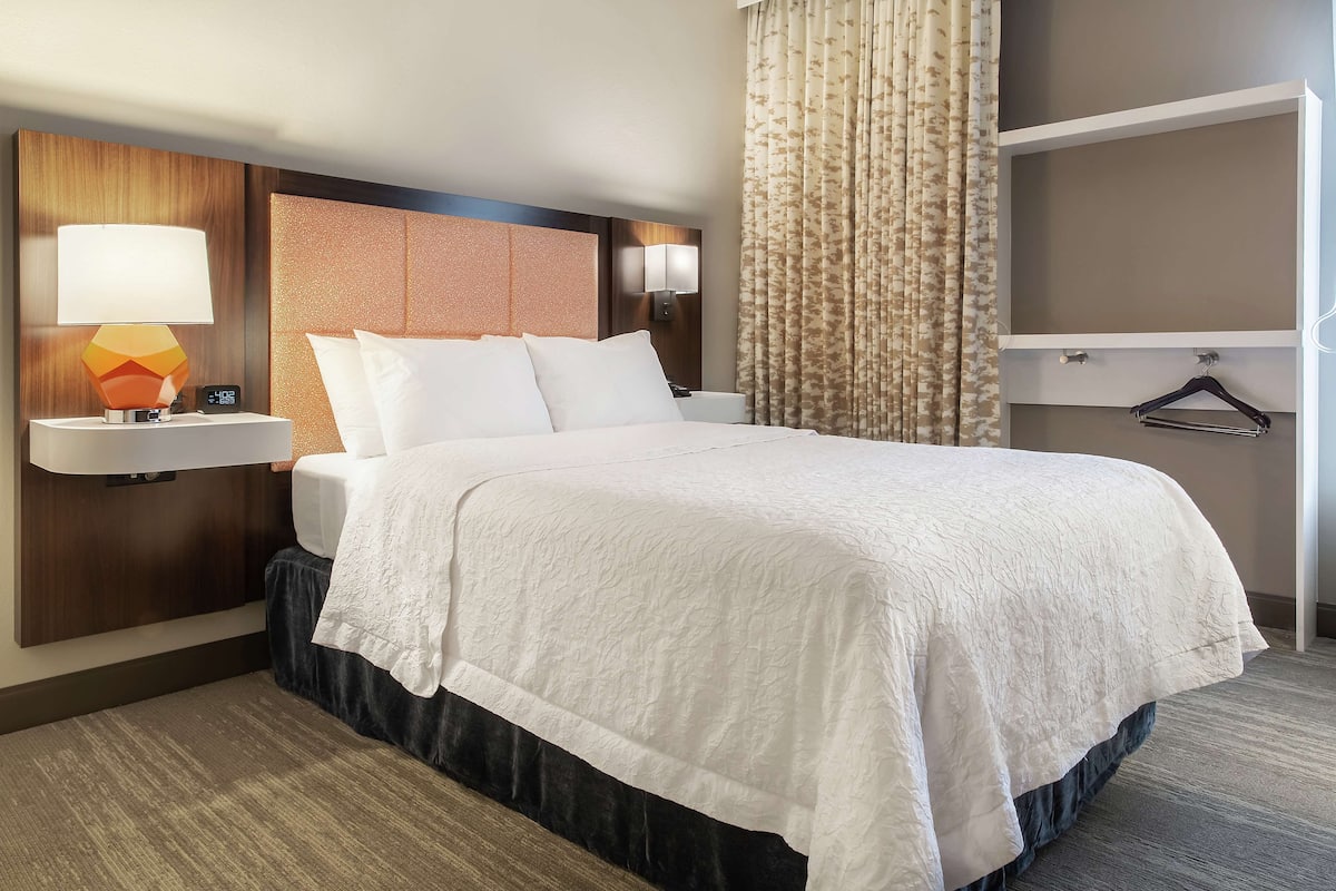 A hotel room with a neatly made bed, a nightstand with a lamp, a wall-mounted shelf, and a hanging coat rack.
