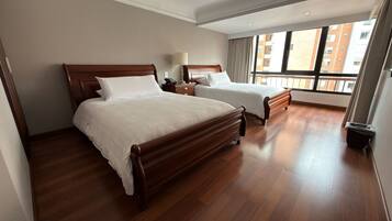 Habitación de hotel con dos camas de madera, una mesita de noche, una lámpara y vistas a edificios a través de amplios ventanales.
