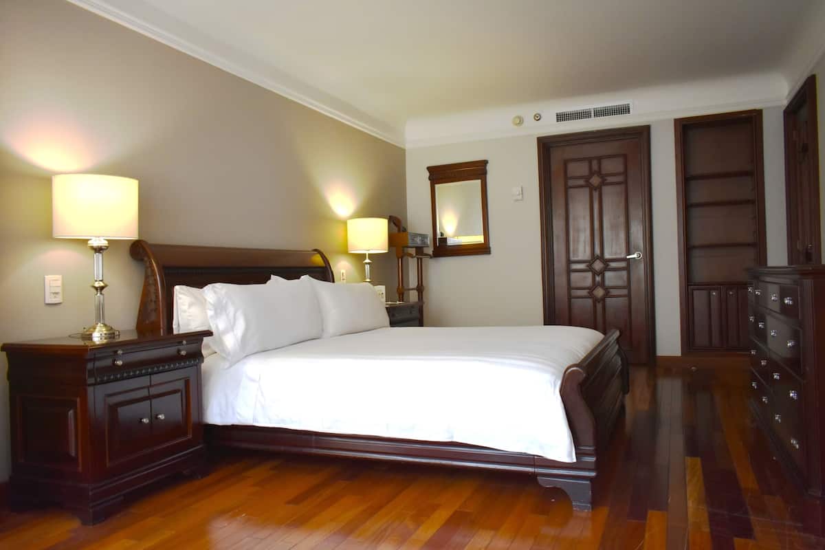 A neatly made bed with white linens, a wooden nightstand with a lamp, a dresser, and a mirror on the wall.