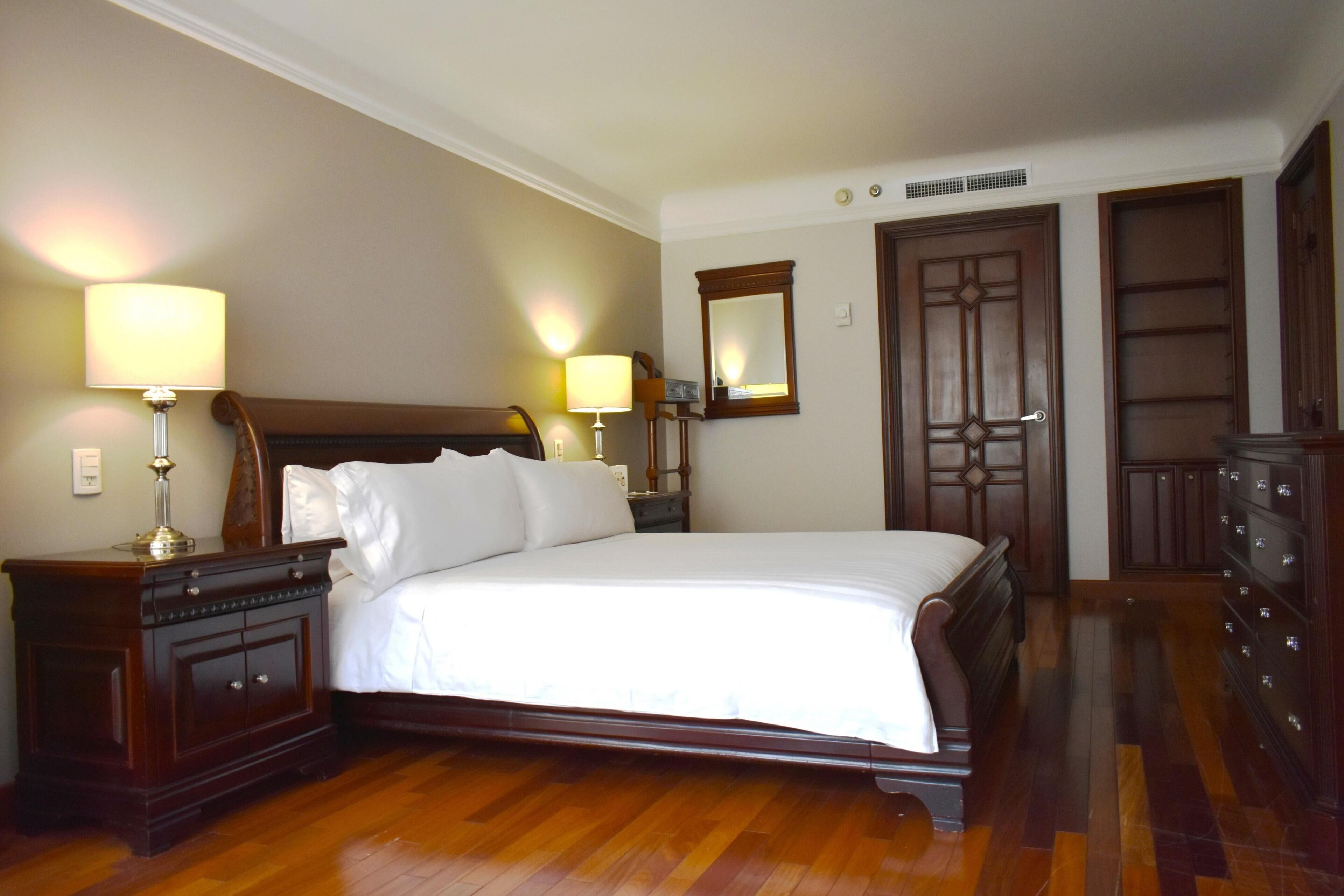 A neatly made bed in a hotel room with wooden furniture, a nightstand with a lamp, a dresser, and a mirror on the wall.