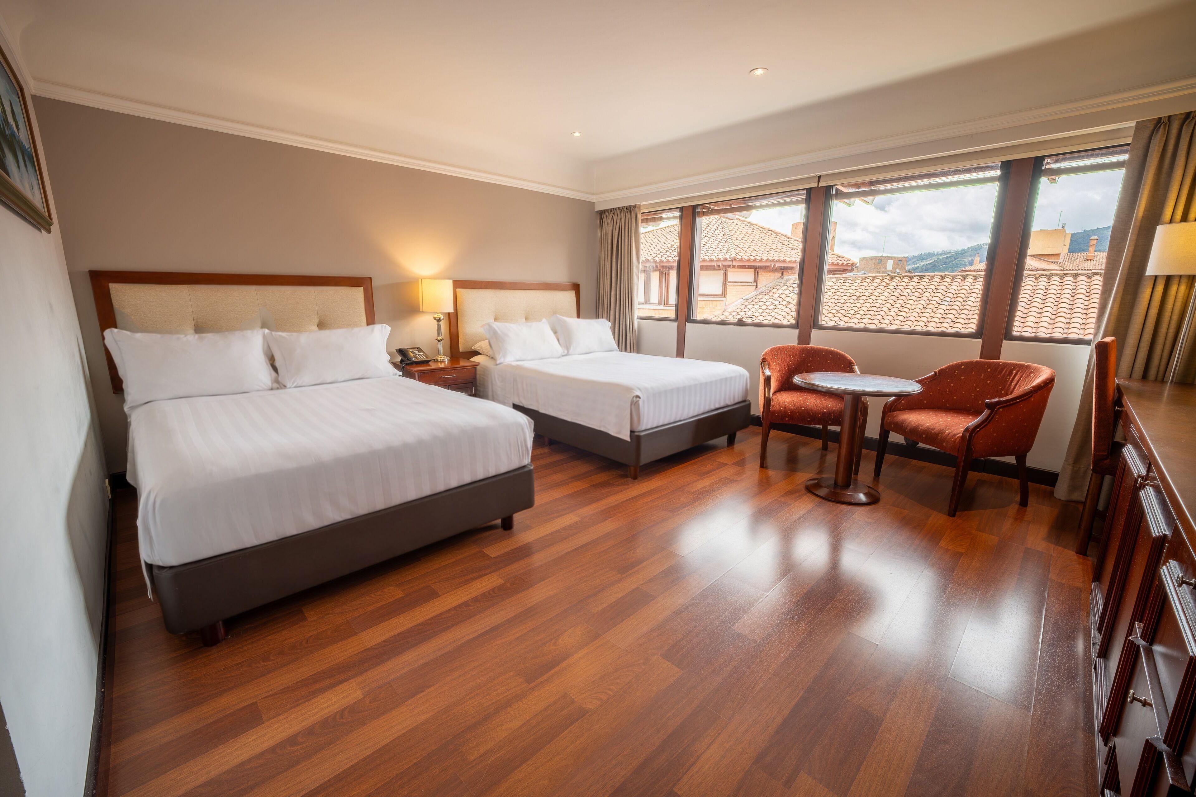 A hotel room with two beds, a small table, a chair, and a view of the cityscape.