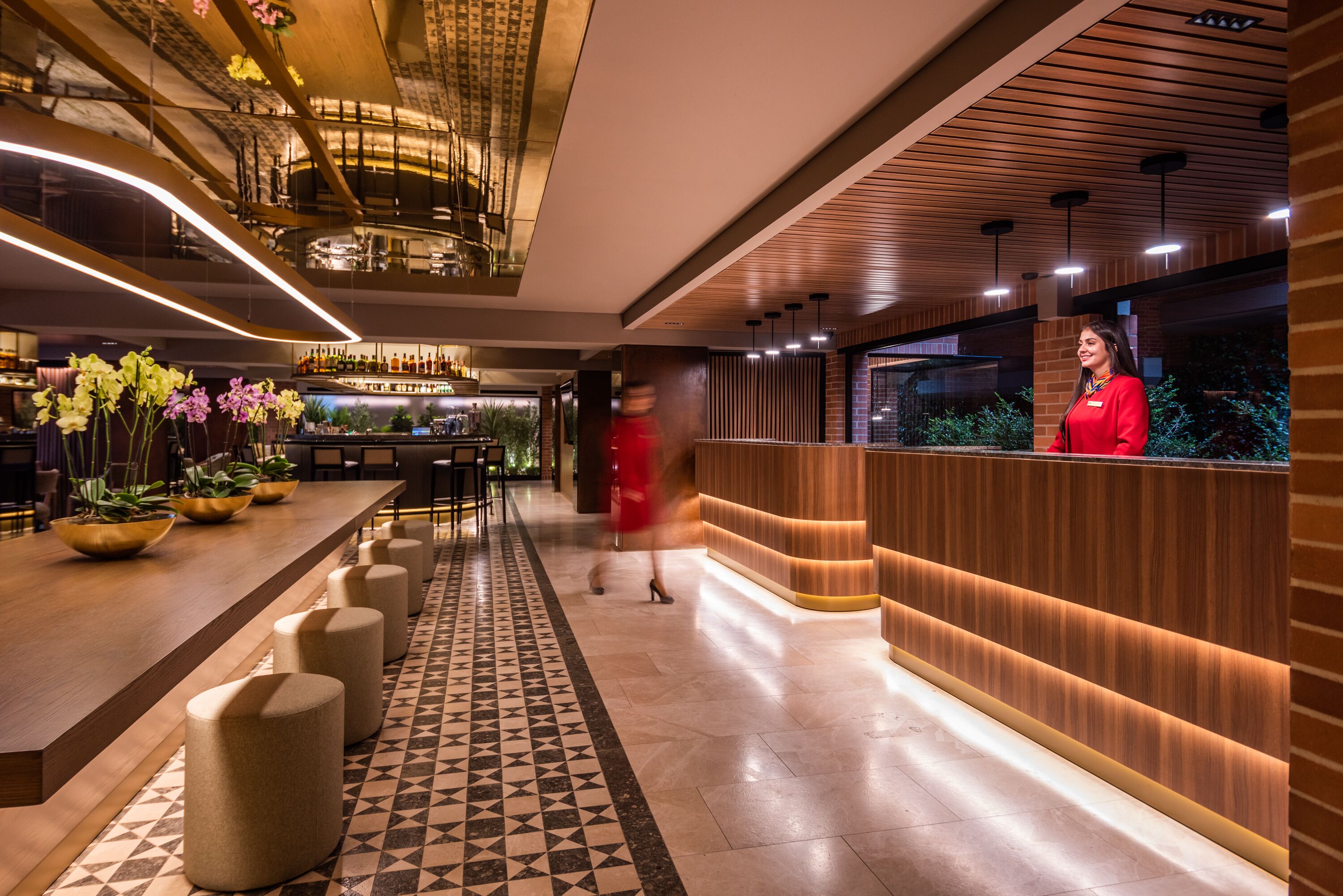 A modern hotel lobby with a long reception desk, a patterned floor, and a bar area with plants and bottles.