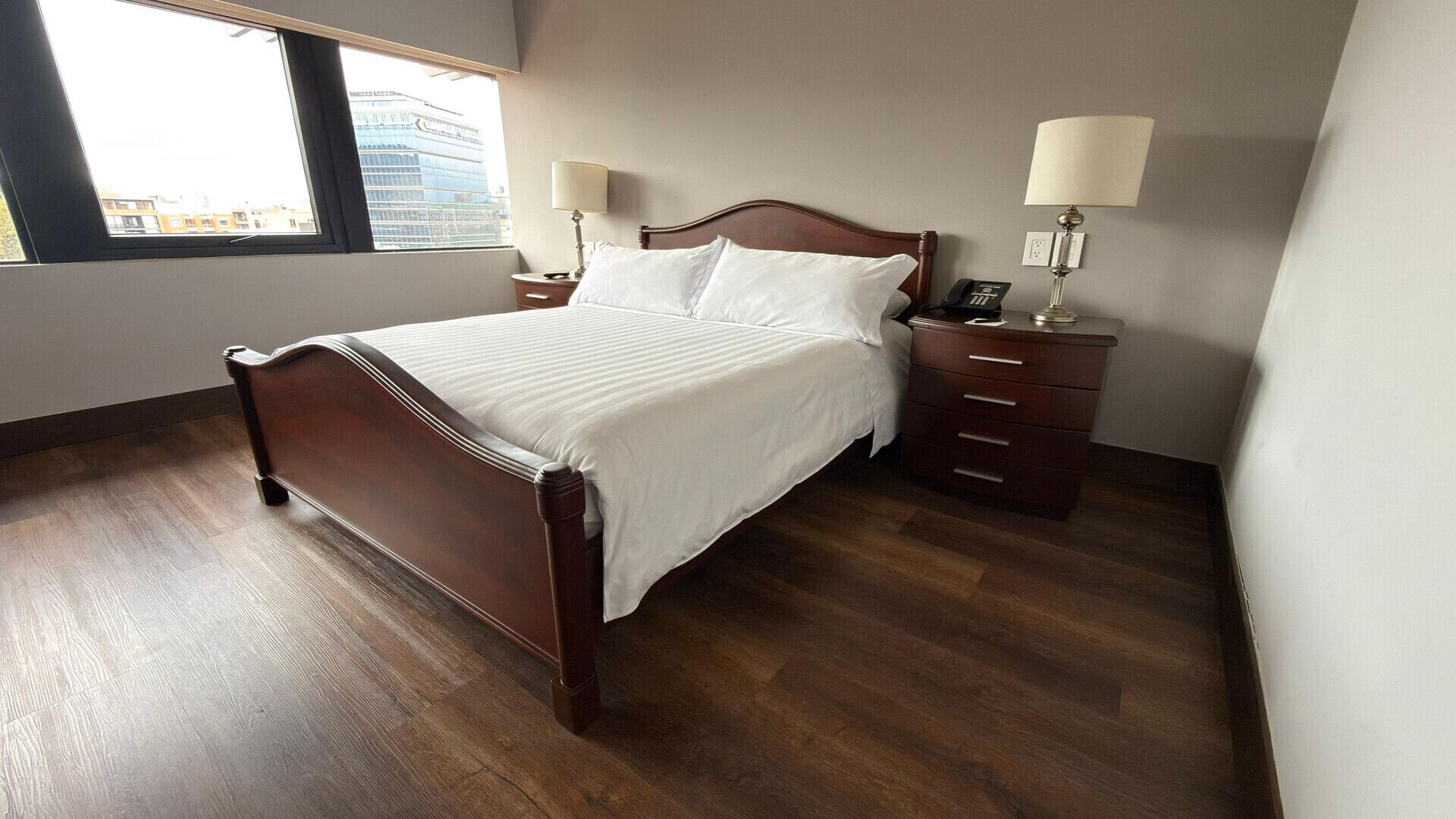 A hotel room with a wooden bed, two bedside tables with lamps, a nightstand, and a window with a view of buildings.