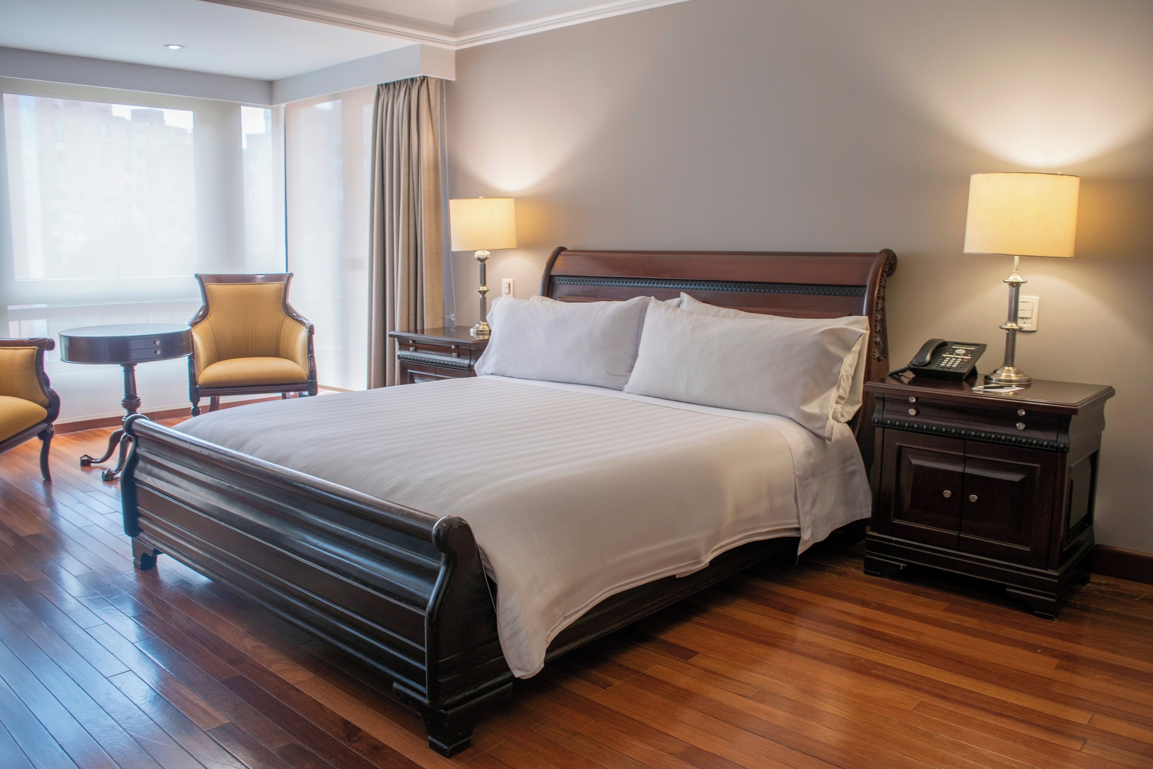 Una cama grande con cabecera de madera, dos mesitas de noche con lámparas, una silla y una ventana con cortinas.