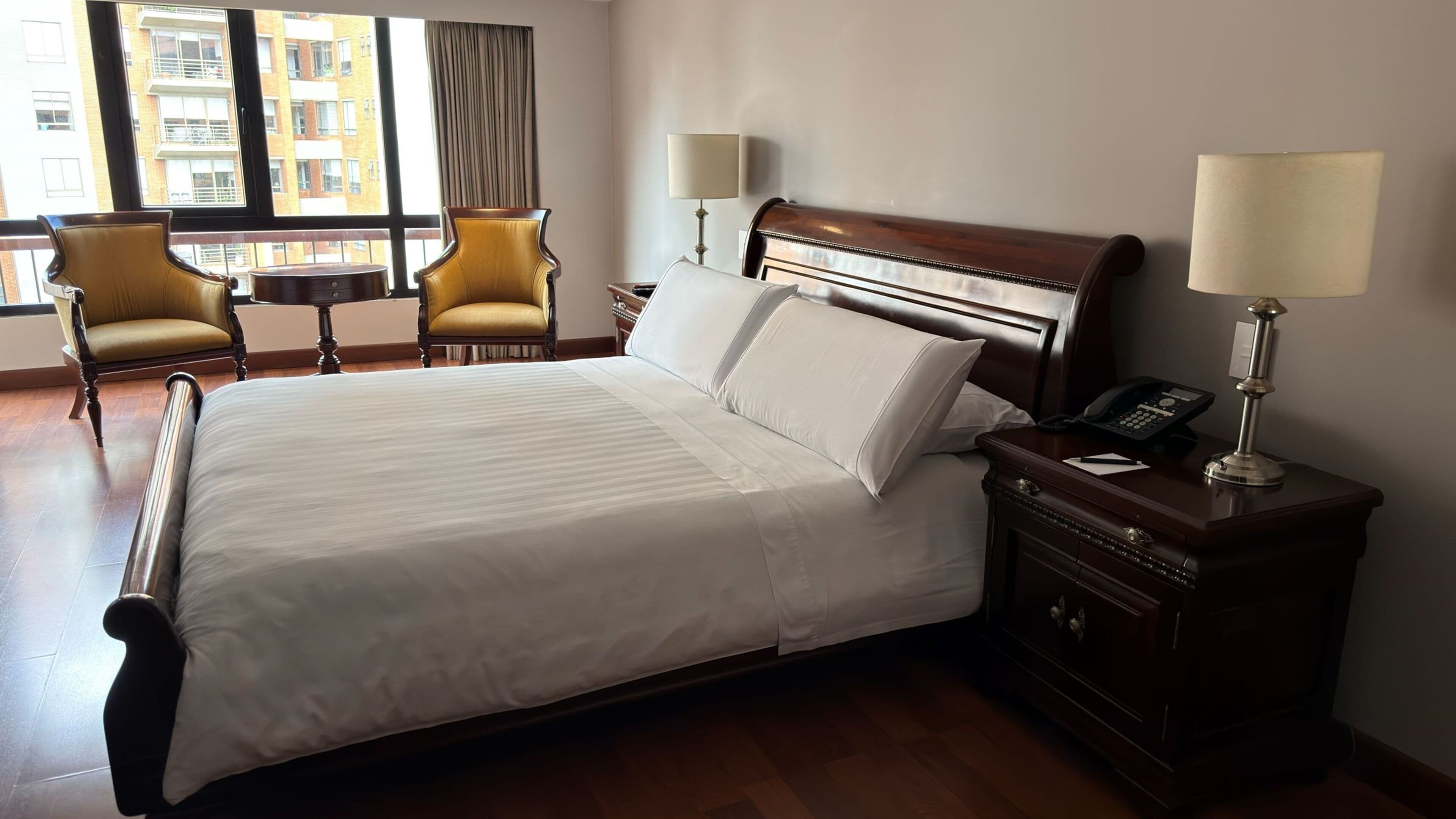 A hotel room with a large bed, two chairs, a nightstand with a lamp, and a view of buildings through the window.