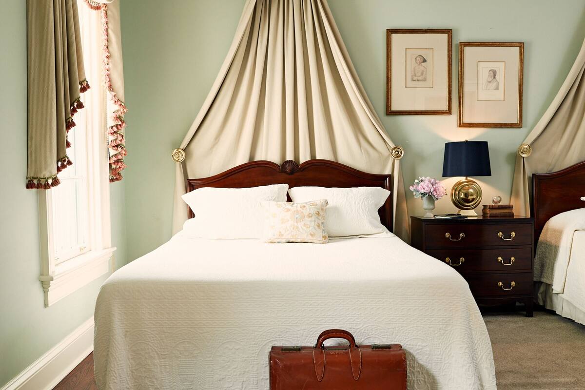 A neatly made bed with a canopy, a bedside table with a lamp, and framed pictures on the wall.