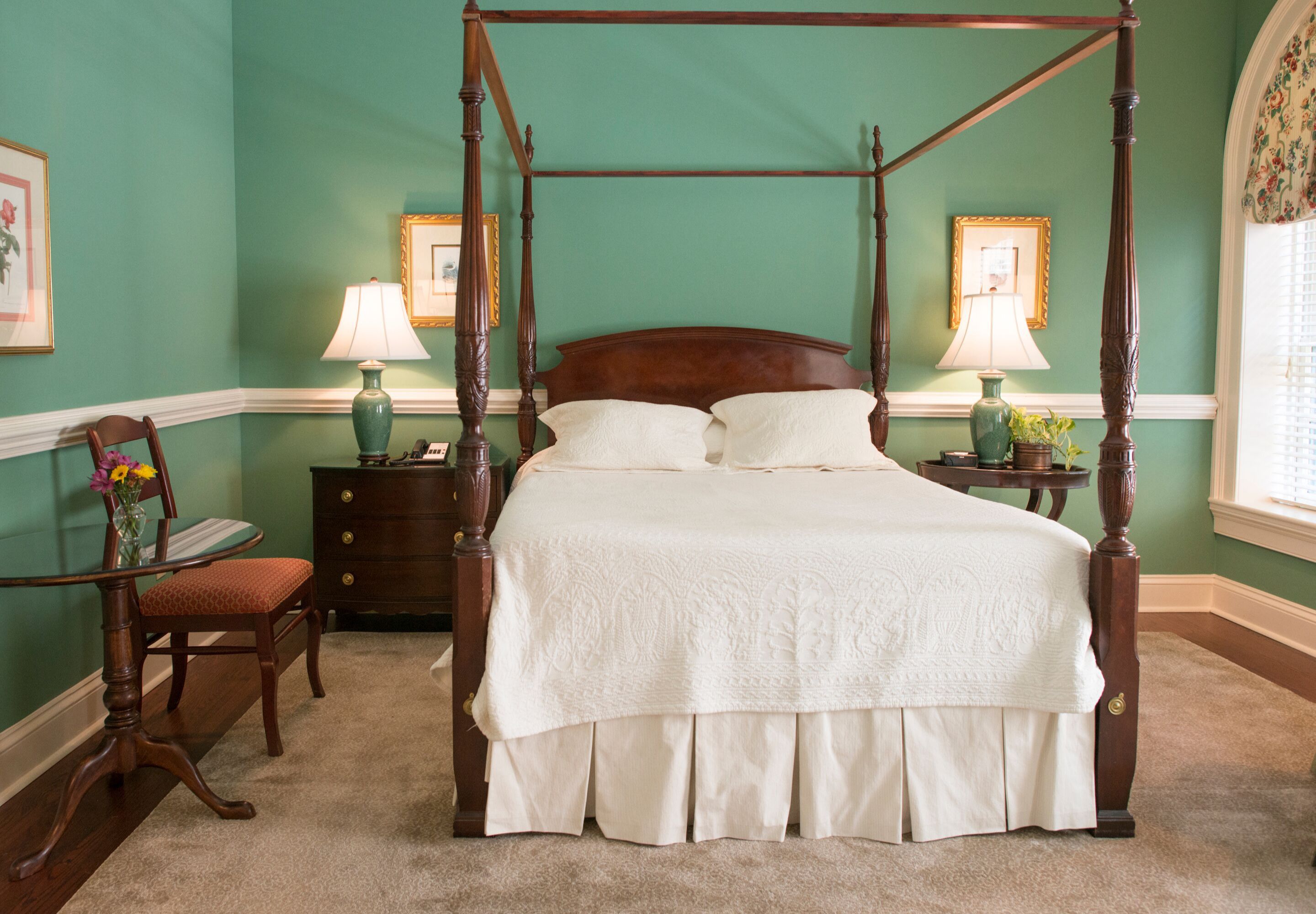 A four-poster bed with a white bedspread, two bedside tables with lamps, a wooden chair, and a vase with flowers.