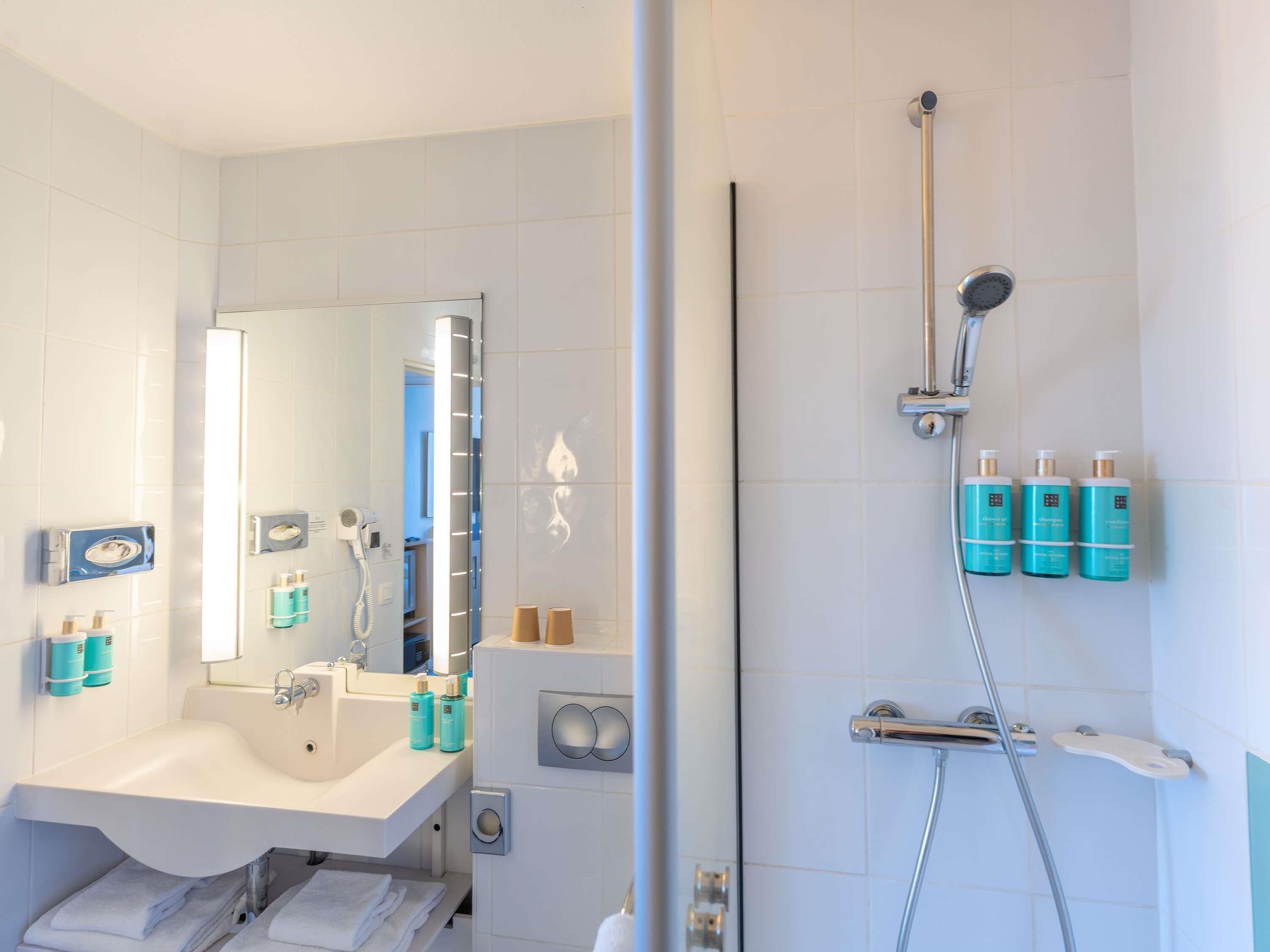 A bathroom with a sink, mirror, towel rack, and a shower area with a handheld showerhead and bottles of shampoo.