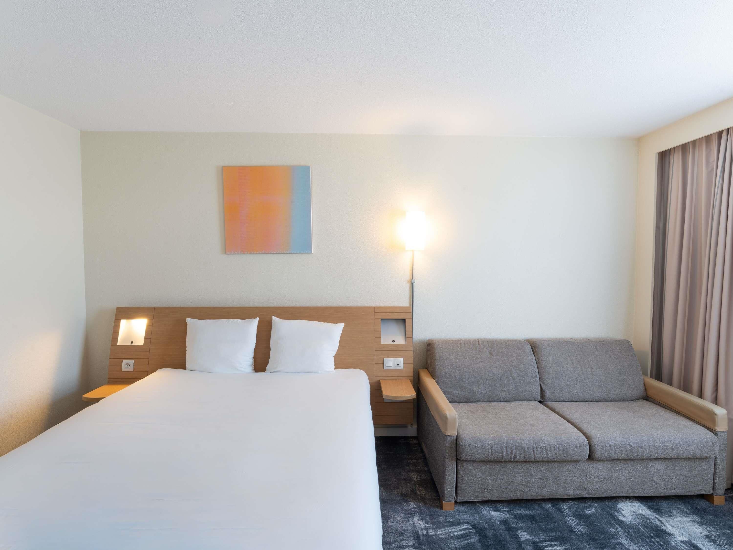 A hotel room with a large bed, a sofa, a nightstand, and a wall-mounted lamp.