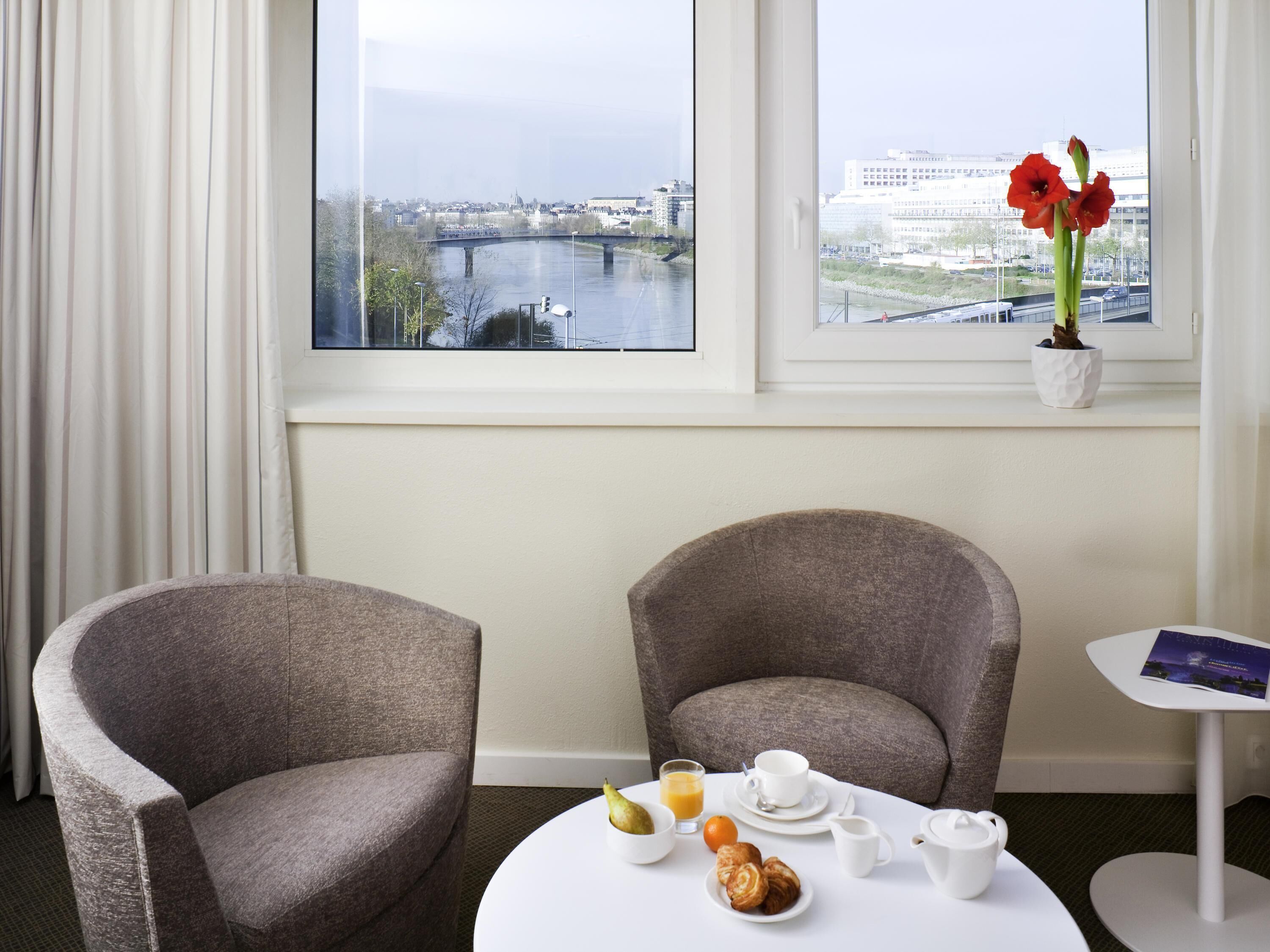 Ein Hotelzimmer mit zwei Sesseln, einem kleinen runden Tisch mit Frühstücksgebäck und einem Fenster mit Blick auf die Stadt und Blumen.