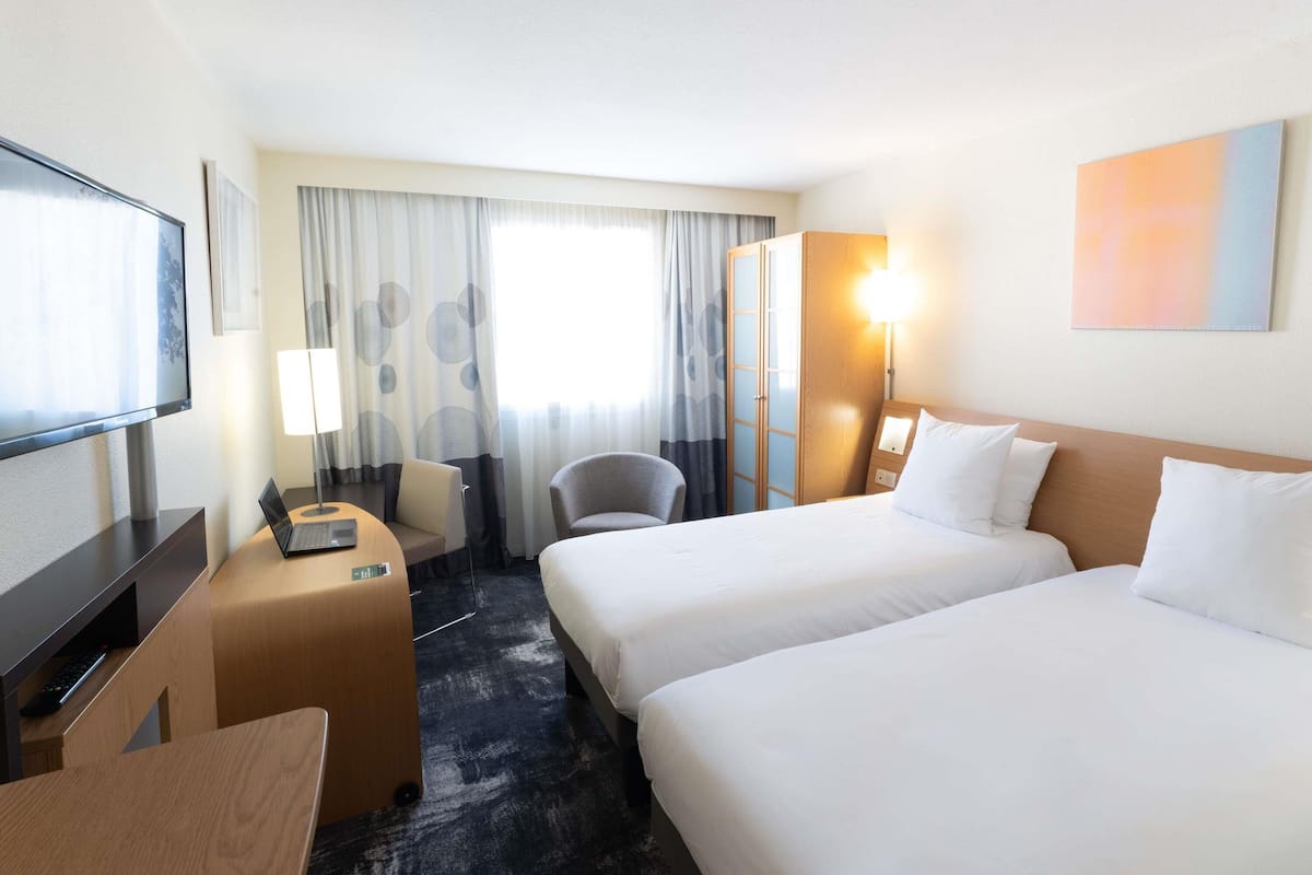 A hotel room with two beds, a desk with a computer, a chair, a TV, and a window with curtains.