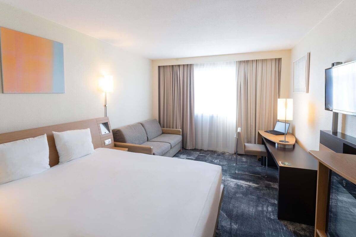 A hotel room with a bed, a sofa, a desk with a laptop, a TV, and a large window with curtains.