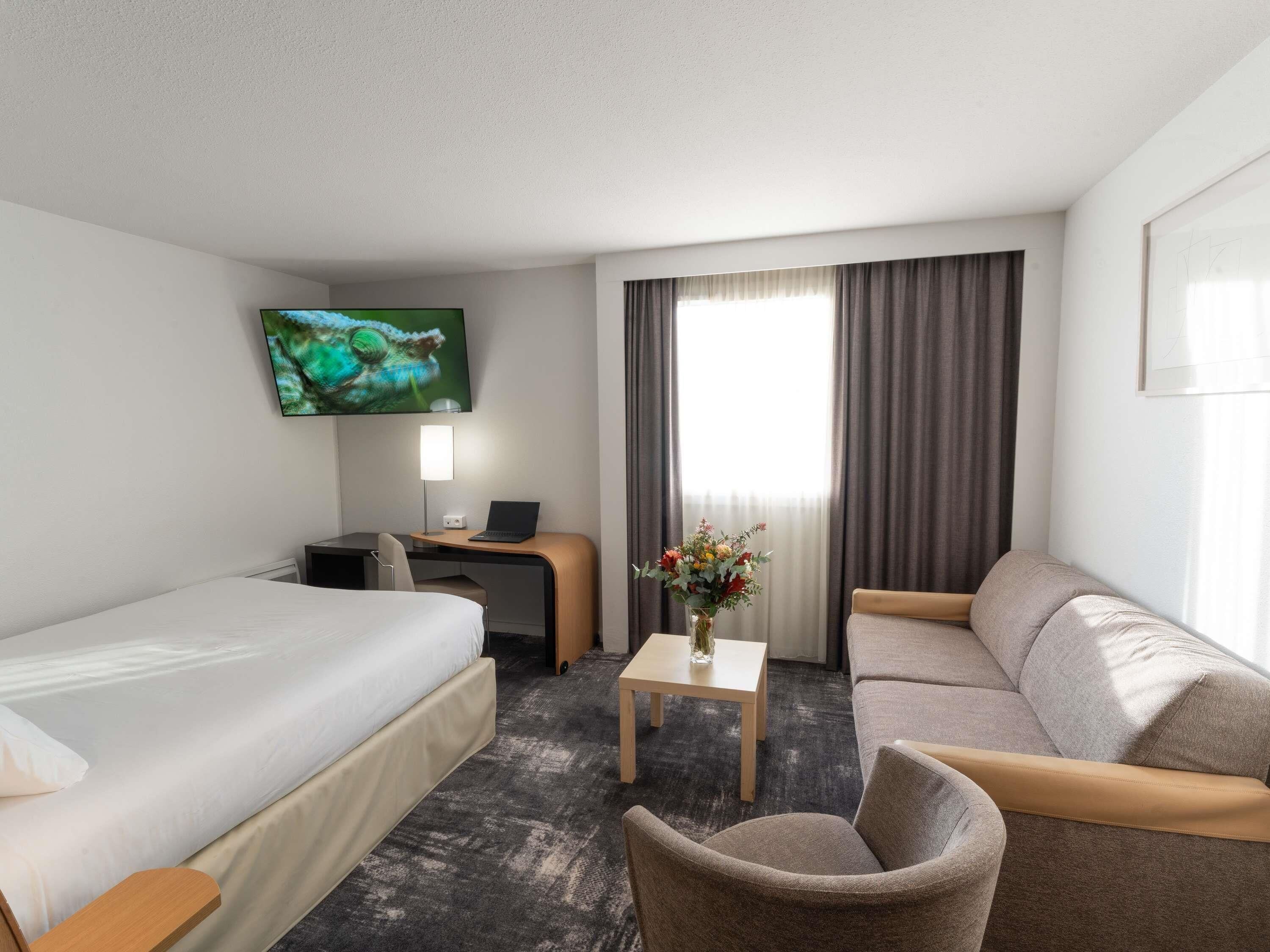 A hotel room with a bed, a desk with a laptop, a chair, a sofa, a small table with a vase, and a TV on the wall.