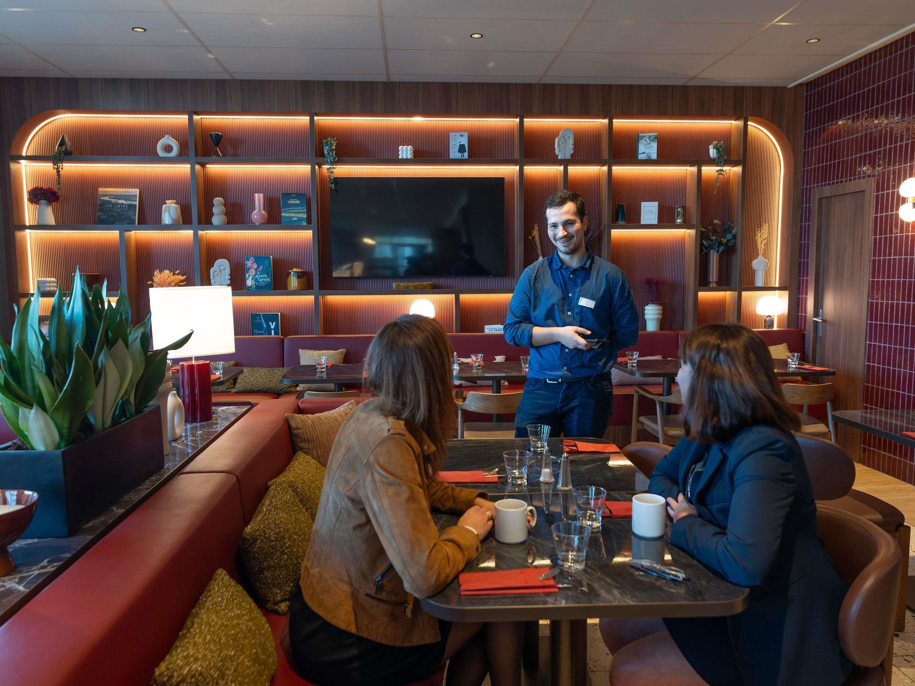 A restaurant with a modern interior, featuring red cushioned seating, a central table with a waiter, and a TV mounted on the wall.