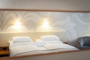 Uma cama bem arrumada com lençóis e almofadas brancos, uma cabeceira com duas luminárias fixadas na parede e uma poltrona cinza.
