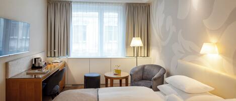Quarto de hotel com uma cama, uma escrivaninha, uma cadeira, uma mesinha e uma televisão.
