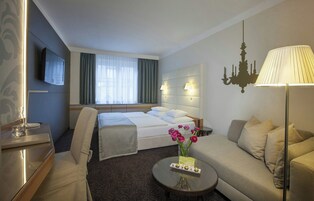 Quarto de hotel com cama, sofá, mesa de centro, televisão e lustre.