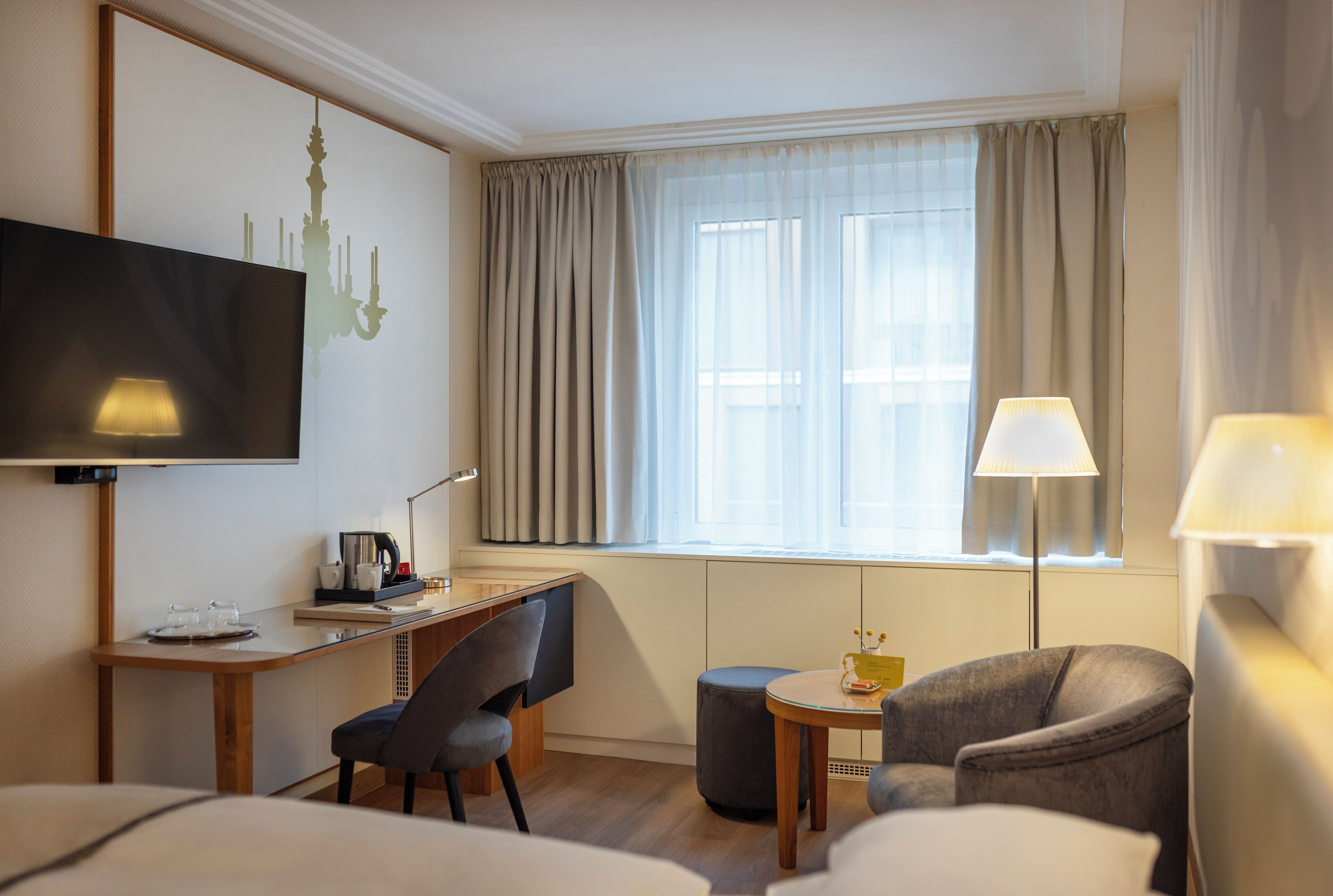 A hotel room with a desk, two chairs, a TV, a lamp, and a window with curtains.