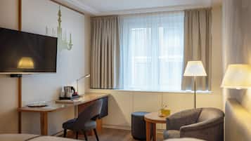 Quarto de hotel com escrivaninha, duas cadeiras, televisão, candeeiro e janela com cortinas.